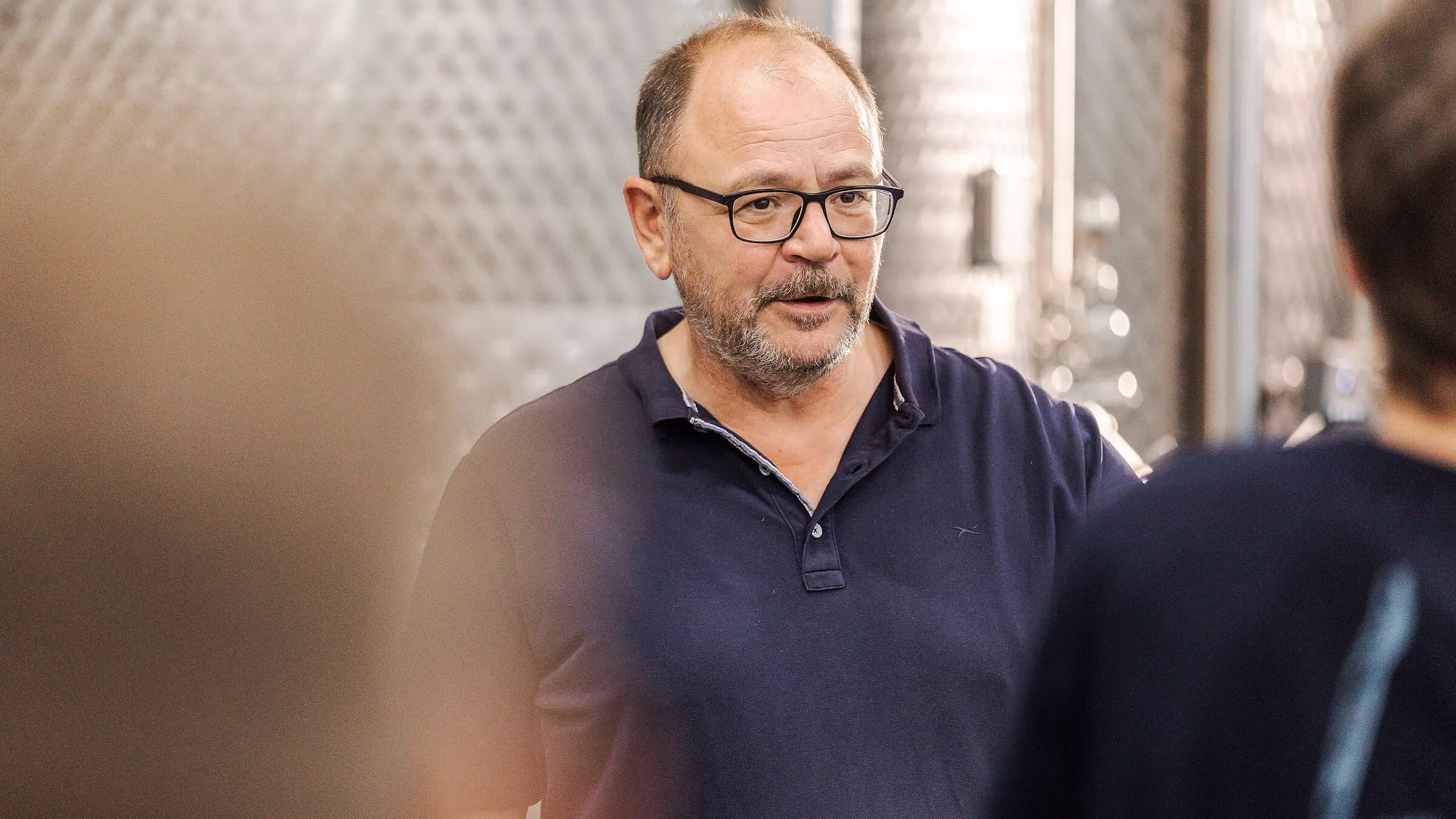 Ein Mann mittleren Alters mit Brille und Bart, der ein marineblaues Poloshirt trägt, steht in einem Haus und scheint zu einer Gruppe zu sprechen. Im Hintergrund ist eine metallische Industriekulisse zu sehen.