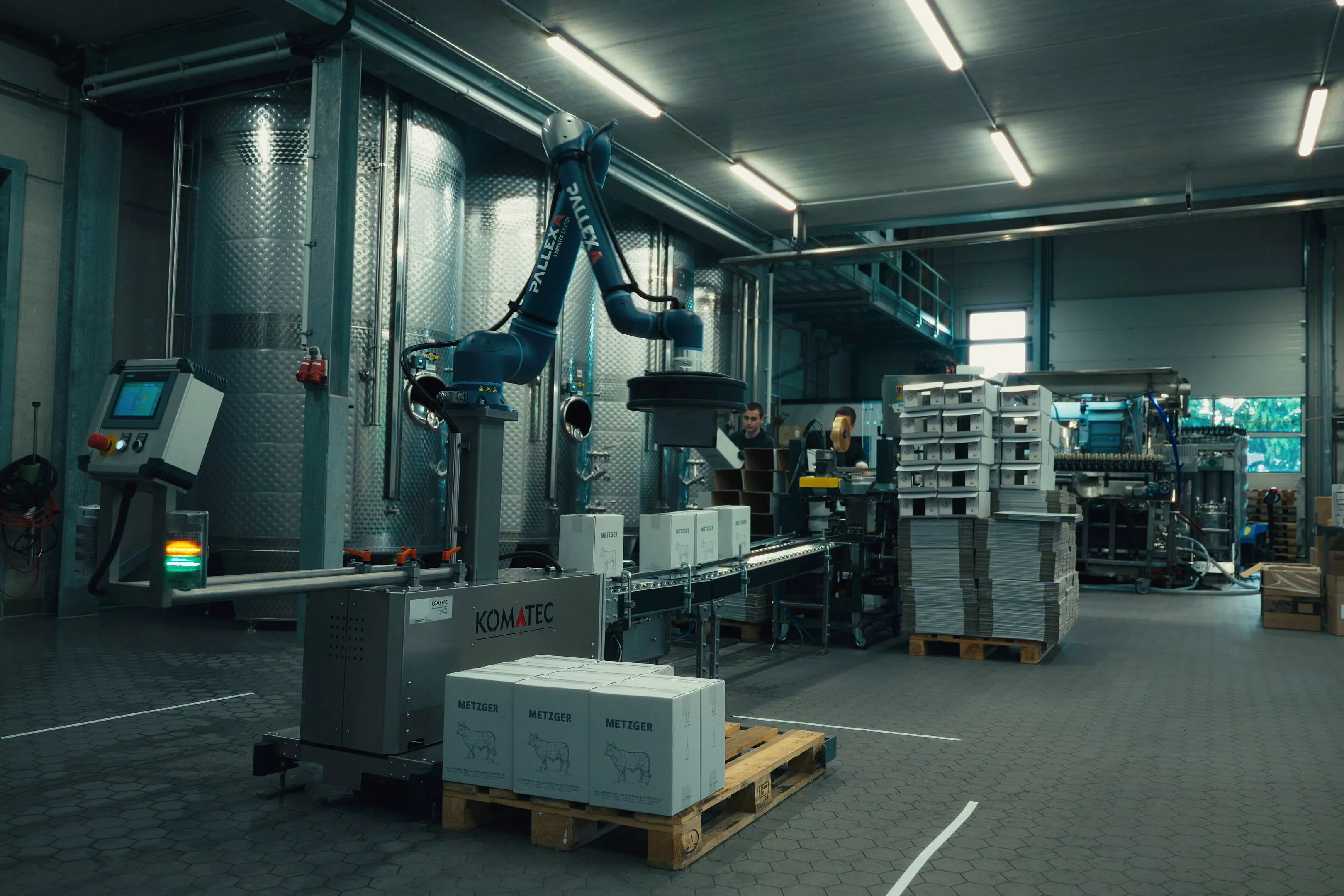 Ein Roboterarm in einer Industrieanlage befördert Kisten auf ein Förderband, das von Metalltanks, gestapelten Tabletts und verschiedenen Geräten umgeben ist.