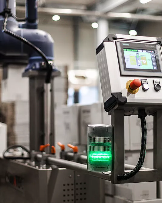 Nahaufnahme einer kompakten Industrie-Steuertafel mit Touchscreen-Display, Not-Aus-Taster und grüner Kontrollleuchte in einer modernen Fabrikumgebung mit unscharfen Maschinen im Hintergrund.