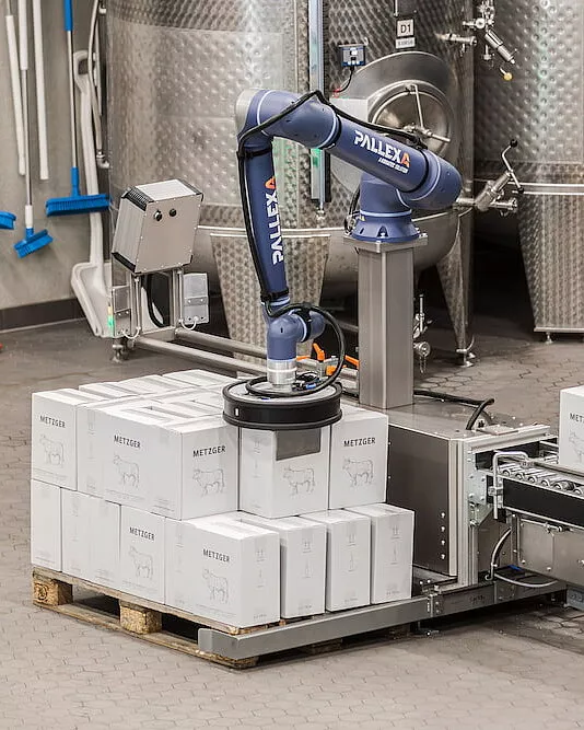 Ein kompakter Roboterarm mit der Aufschrift "PALLEXA" palettiert sicher weiße Kisten mit der Aufschrift "METZGER" auf einer Holzpalette in einer Industrieumgebung mit Metalltanks und Rohren im Hintergrund.