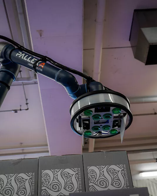 A robotic arm called "PALLEXA" palletizes and lifts a crate with a suction gripper in an industrial environment with compactly stacked patterned crates at the bottom and a ceiling with exposed pipes at the top.