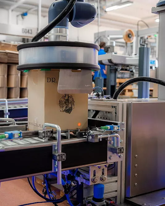 Ein kompakter Roboterarm mit Saugvorrichtung hebt einen Karton sicher von einem Förderband für sicheres Palettieren in einem Industrielager, umgeben von Maschinen und Geräten.
