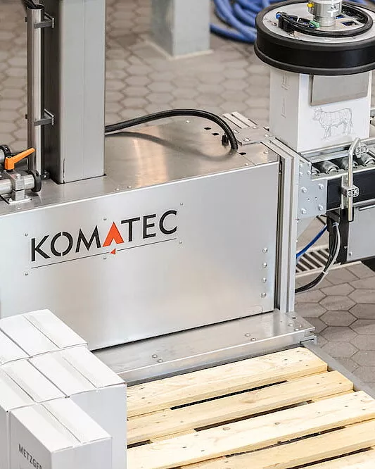 Eine Maschine mit der Aufschrift KOMATEC verarbeitet Kartonagen auf einem Fördersystem. Im Vordergrund sind mehrere weiße Kartons auf einer Holzpalette gestapelt. Die Maschine steht auf einem grauen Fliesenboden.
