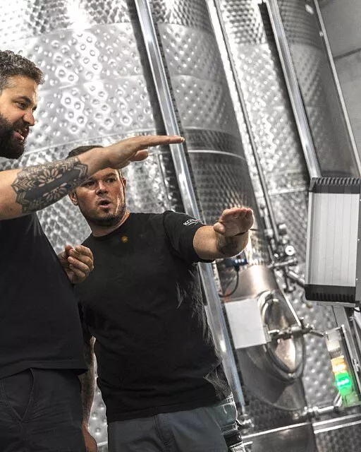 Zwei Männer in schwarzen T-Shirts stehen vor großen Metalltanks und diskutieren etwas, während sie mit Gesten auf einen Monitor zeigen.